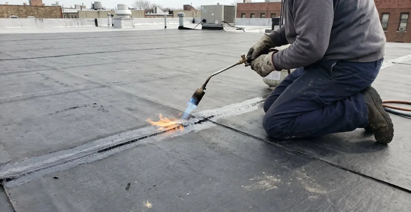 Couvreur professionnel utilisant un chalumeau à propane pour souder les joints d'une membrane élastomère noire, vu par-dessus l'épaule avec focus sur la flamme bleue et la surface de la membrane